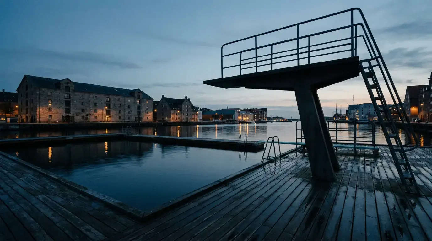 A Nordic harbour bath at dusk: a tall steel diving tower over still black water, timber decking, and lit warehouse buildings along the far quay.