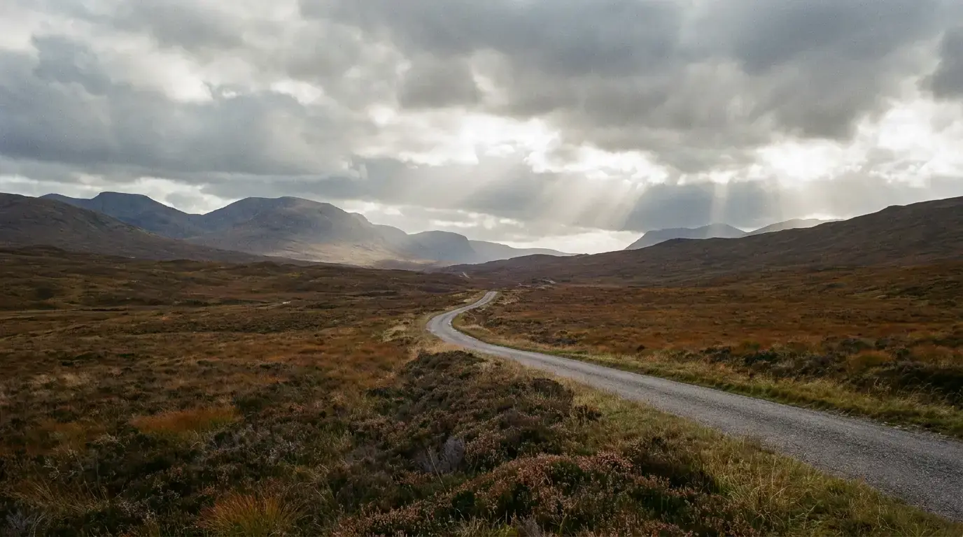 An empty single-track road winding away through Highland moorland toward distant hills, shafts of sun breaking through heavy cloud.